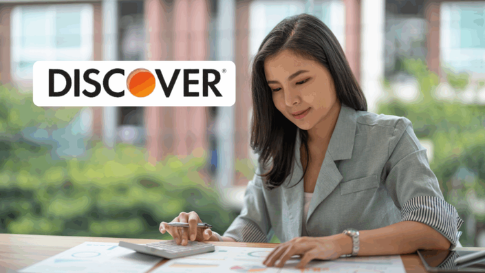 A focused young woman is using a calculator and reviewing documents at a desk with a blurred outdoor background. The Discover logo is digitally overlaid in the top left corner.