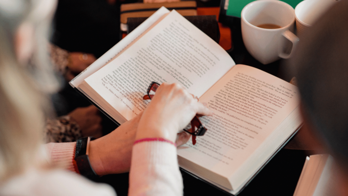 Close-up of a person reading an open book, holding eyeglasses in one hand, with cups of coffee and other books around, creating a cozy reading environment focused on Books of Personal Finance.