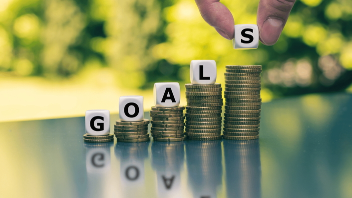 A series of stacked coins arranged in ascending order, with each stack supporting a white cube displaying a letter, spelling out the word "GOALS." A hand is placing the final cube with the letter "S" on the tallest stack. The background is blurred with greenery, symbolizing financial growth and goal-setting.