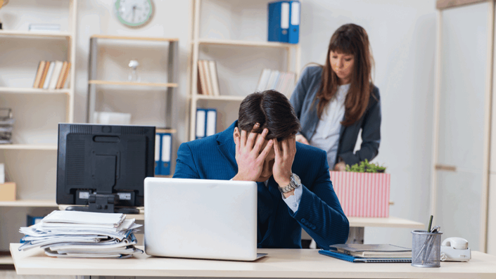 An office worker sits at a desk with head in hands, looking distressed, while a colleague in the background packs a box, illustrating the theme of job displacement due to AI and Automation.