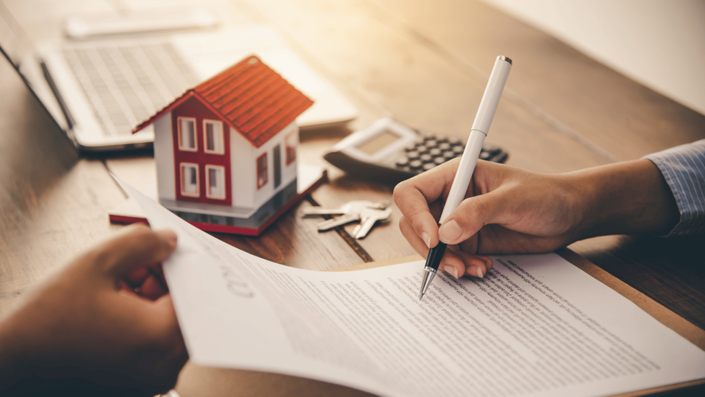 A person signing a mortgage contract with a pen on a wooden table. A small model house, keys, a calculator, and a laptop are placed nearby, symbolizing homeownership and mortgages.