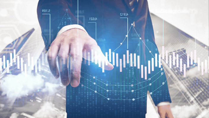 A businessman in a suit points his finger towards a holographic display of financial charts and graphs, with blurred city buildings in the background, representing the digital future of finance and how money can be managed with a robo-advisor.